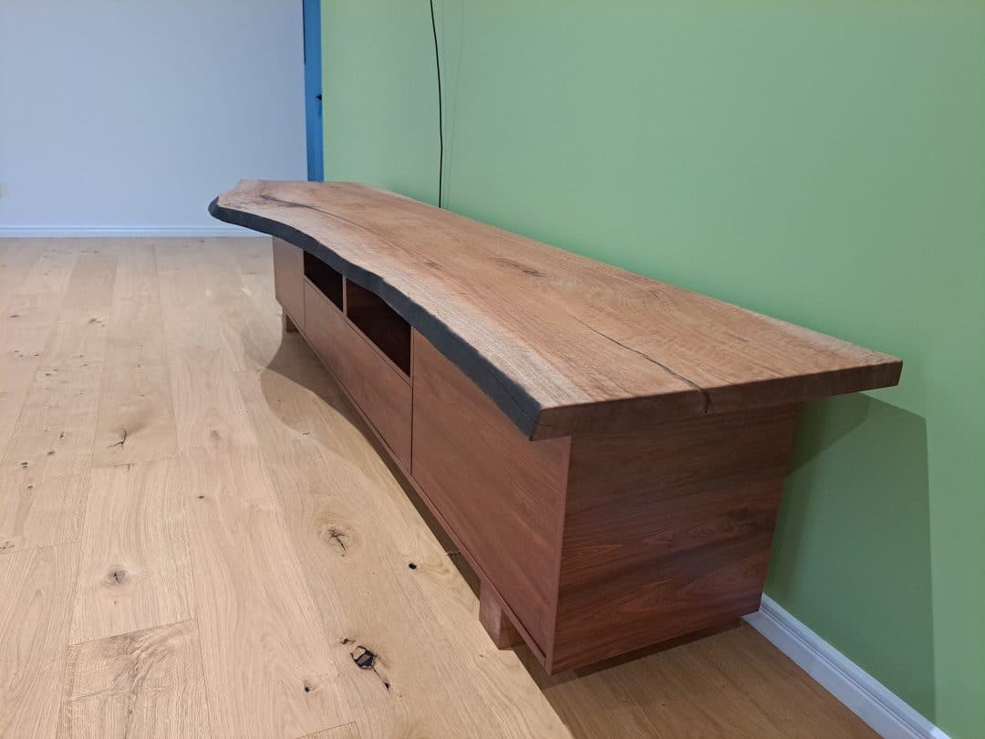 A wooden sideboard with a natural, unpolished live edge top rests against a green wall in a room with light hardwood flooring. The sideboard features an open shelf and cabinets, showcasing the natural grain and texture of the wood.