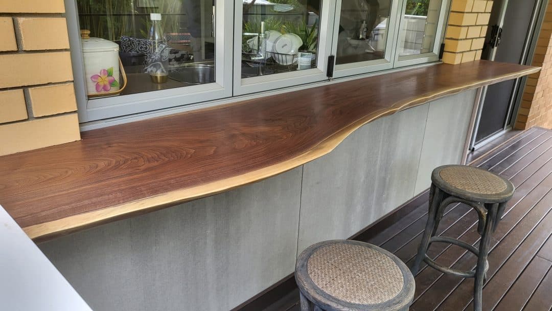 A sleek wooden bar counter with a natural, wavy edge extends along an outdoor patio area. Bar stools with woven seats are positioned in front of it. The background reveals a window peeking into a cozy kitchen space with potted plants and kitchenware.