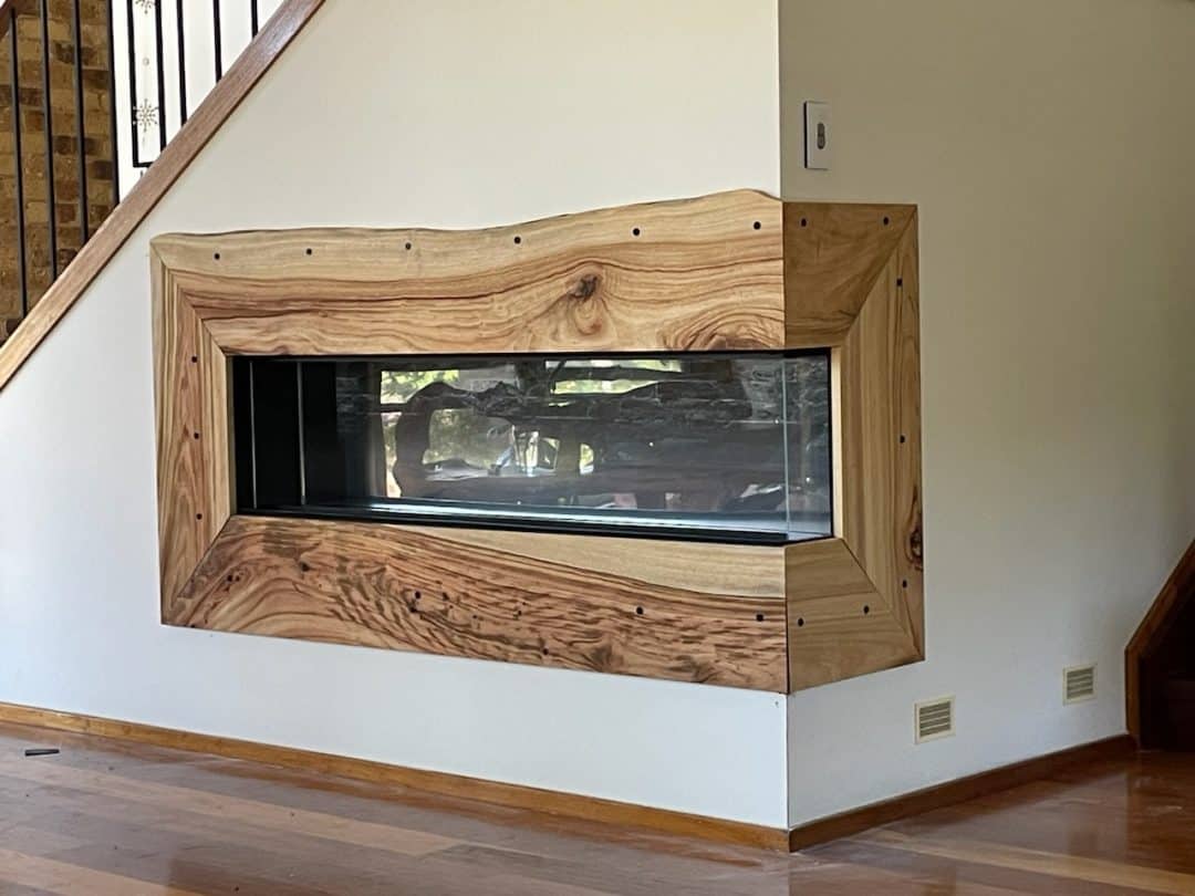 A modern fireplace recessed into the wall, featuring a large wooden frame with natural grain and texture. The fireplace borders two rooms, visible through the glass on both sides. It is surrounded by a white wall and next to a wooden staircase with a wrought iron railing.