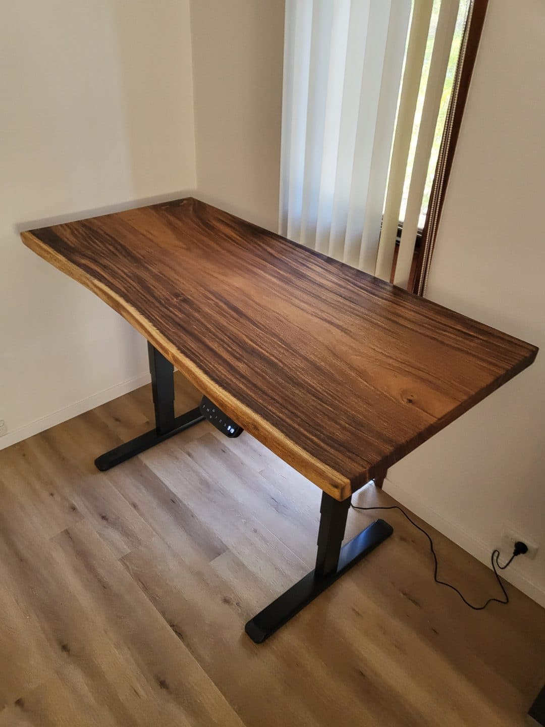 A wooden desk with a natural live edge sits in the corner of a room with light-colored walls and floor. The desk has black metal legs and is placed near a window with vertical blinds. An electrical cord is visible underneath.