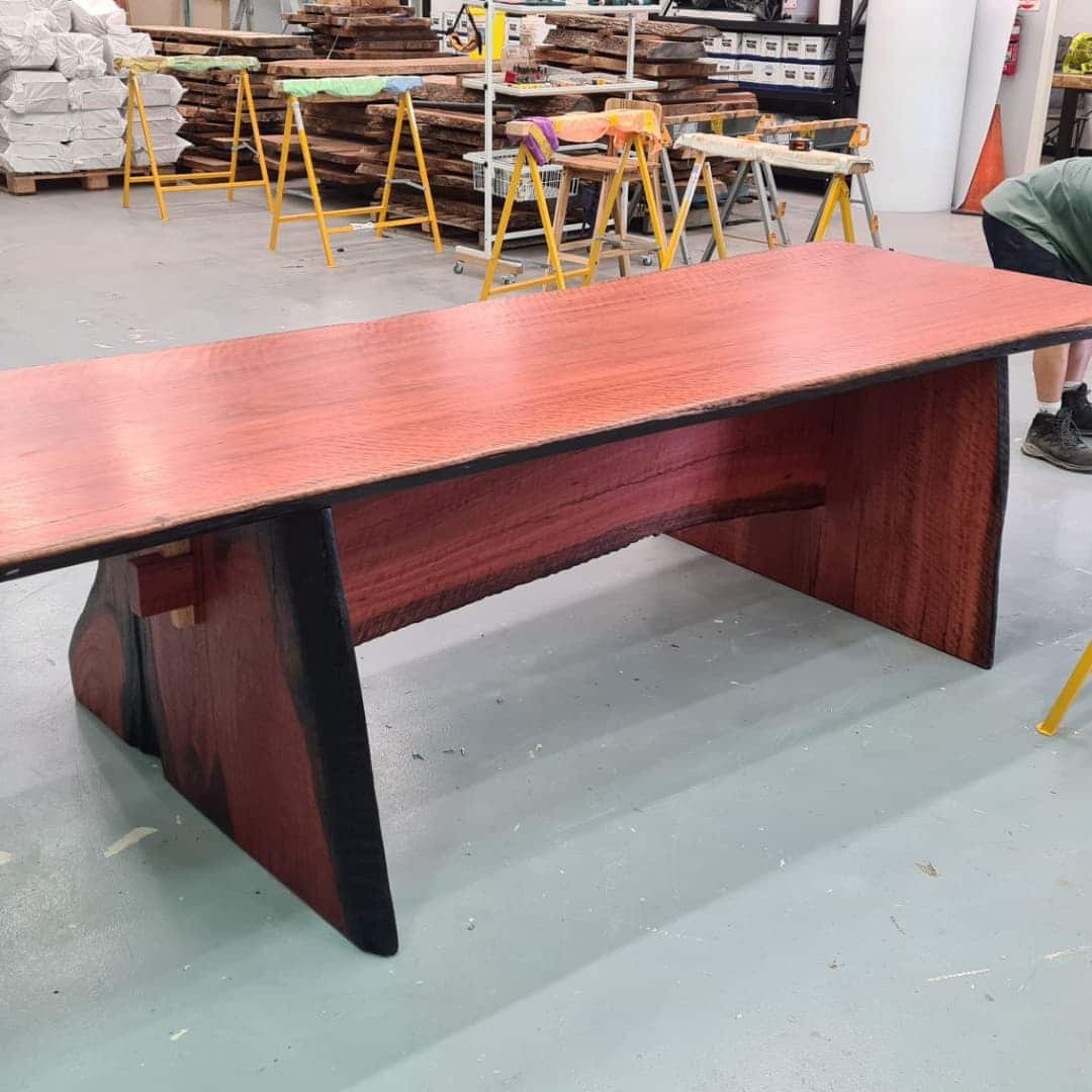 A large wooden table with a reddish-brown top and black-edged sides stands in a workshop. Stacks of wood and industrial tools can be seen in the background, and a person is partially visible to the right.