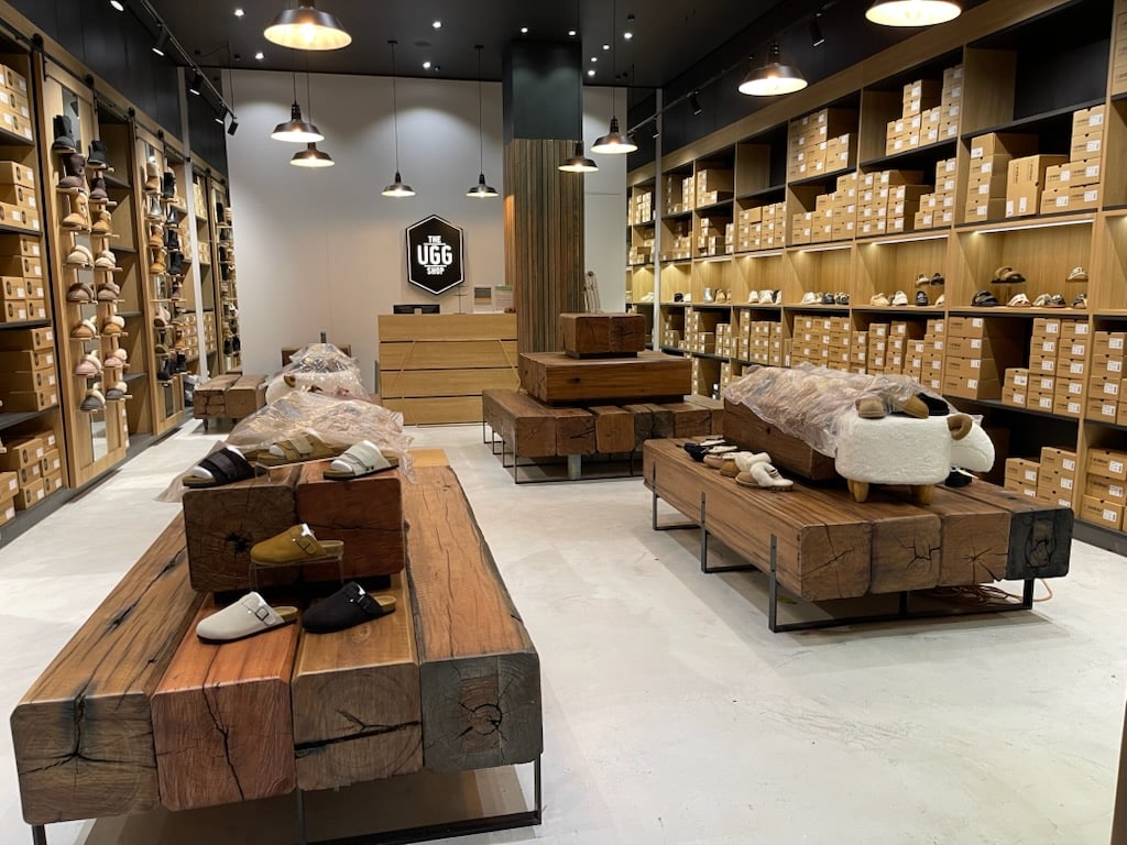 A modern shoe store with wooden tables displaying various slippers and shoes, shelves filled with shoe boxes, bright lighting, and a UGG sign behind the checkout counter at the back.