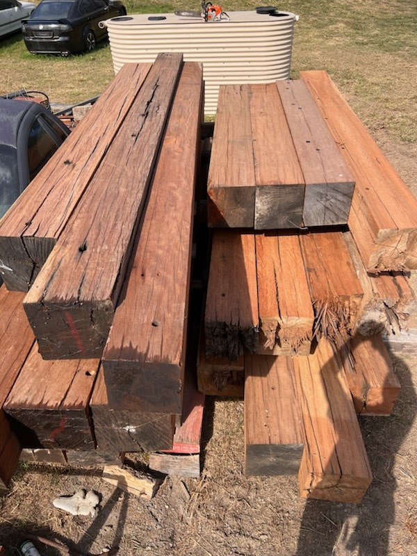 Large, rough-cut wooden beams are stacked outdoors on grass, with visible cracks and weathering. A car and a large water tank are in the background.